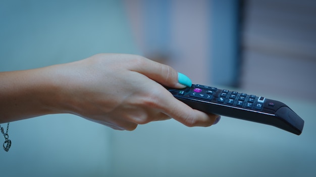 Control remoto de televisión en manos de una mujer apuntando a la televisión y cambiando de canal. Cerca de mujer sosteniendo el controlador y presionando el botón sentado en el sofá frente a la televisión.