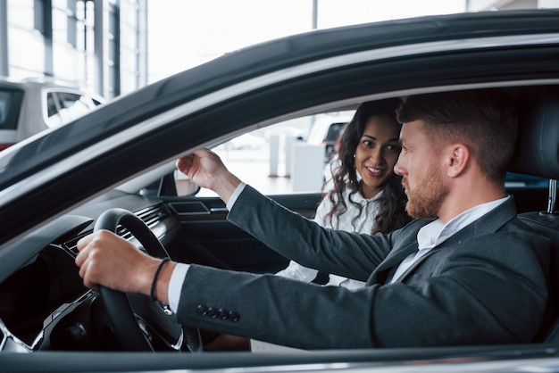 Conversación activa. Encantadora pareja exitosa probando coche nuevo en el salón del automóvil