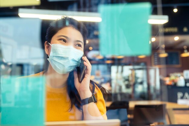 Conversación de protección de máscara facial y ropa casual femenina asiática de agencia creativa con un teléfono inteligente independiente que trabaja con una computadora portátil en el espacio de oficina del área de coworking con frescura con fondo de oficina borroso