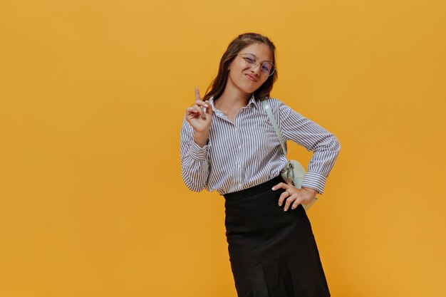 Cool emocional joven apunta hacia arriba sobre fondo naranja aislado Encantador adolescente en uniforme escolar mira a la cámara