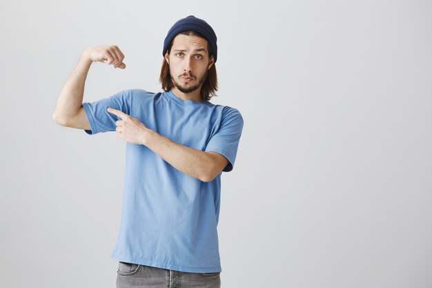 Foto gratuita cool hipster guy en camiseta azul y gorro flex bíceps, presumiendo con músculos fuertes