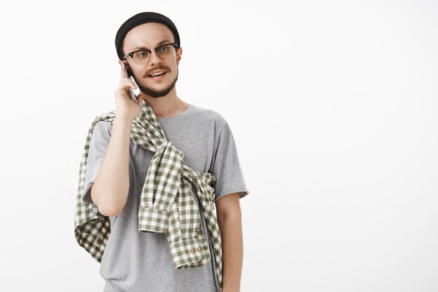 Cool hipster guy llamando a compañeros invitando a venir y agarrar oso sosteniendo el teléfono inteligente cerca de la oreja mientras habla por teléfono mirando a la derecha con expresión normal casual de pie en gorro negro y gafas