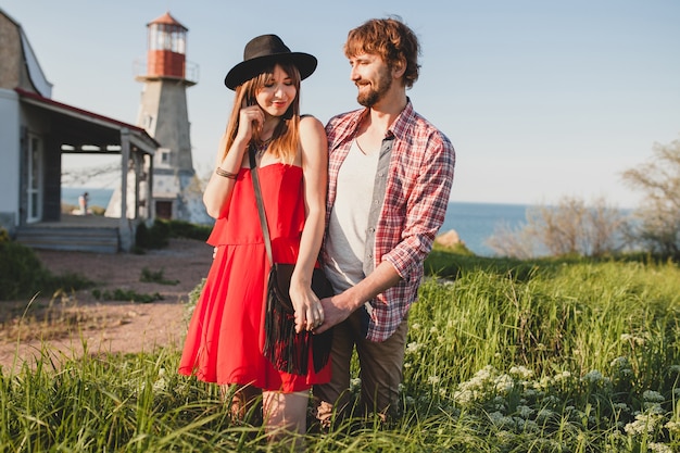 Cool joven elegante pareja enamorada en el campo, estilo bohemio indie hipster, vacaciones de fin de semana, traje de verano, vestido rojo, hierba verde, tomados de la mano, sonriendo