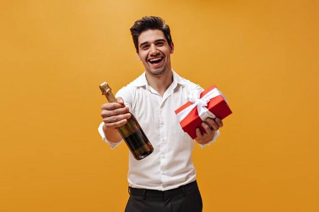 Cool joven emocional con camisa blanca y pantalón negro se regocija, sostiene una caja de regalo roja y una botella de champán en la pared naranja.