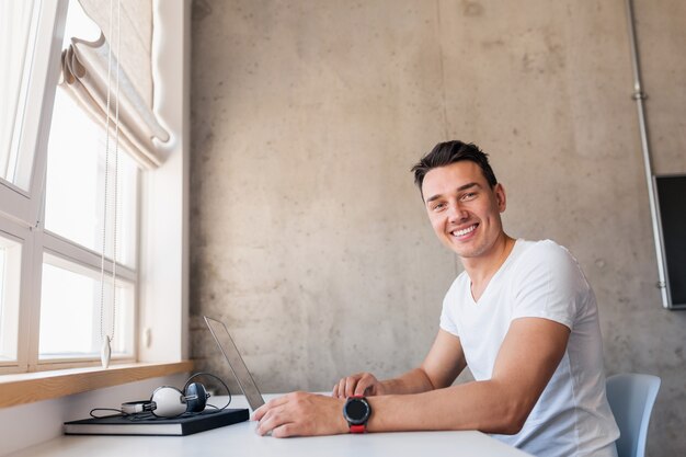 Cool joven guapo sonriente en traje casual sentado en la mesa trabajando en la computadora portátil