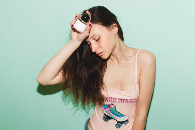 Cool joven hermosa mujer hipster posando contra la pared azul, sosteniendo una botella de perfume de agua de tocador de perfume