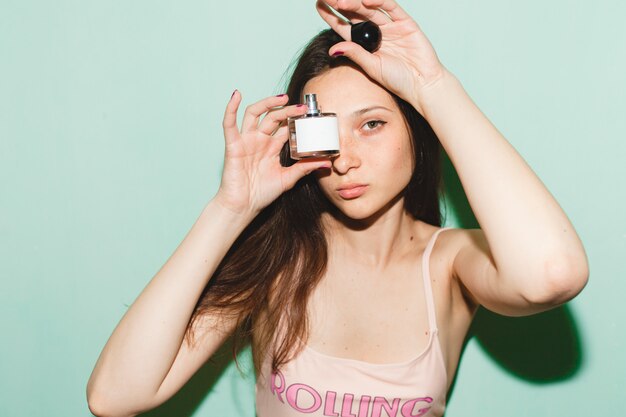 Cool joven hermosa mujer hipster posando contra la pared azul, sosteniendo una botella de perfume de agua de tocador de perfume