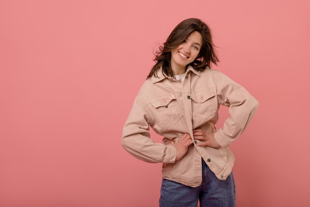 Cool joven morena caucásica en chaqueta mantiene sus manos en la cintura sobre fondo rosa con espacio para texto Concepto de belleza femenina de estilo de vida