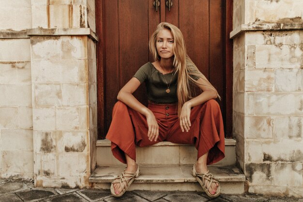 Cool joven rubia triste mujer de pelo largo con pantalones rojos sueltos y camiseta caqui se sienta en pose divertida cerca de la puerta de madera y mira al frente