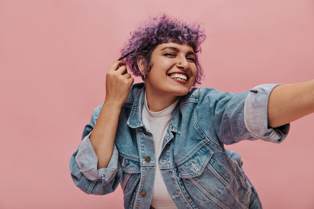 Cool mujer con piercings y cabello morado sonríe y toma selfie. Mujer elegante en camiseta blanca posando en rosa.