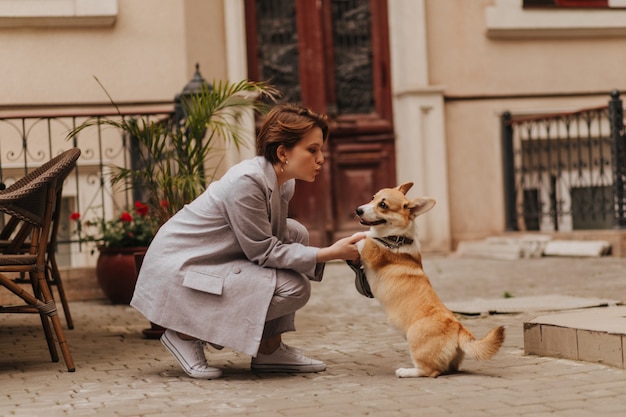 Cool mujer en traje gris juega con corgi afuera. Chica atractiva en chaqueta y pantalones de gran tamaño abrazando a su perro