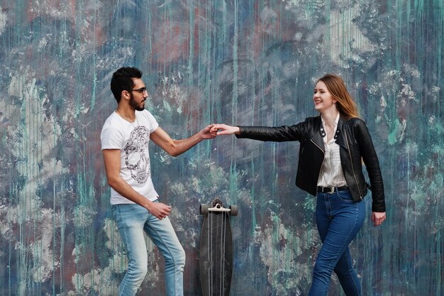 Cool pareja multirracial posando contra la pared con longboard