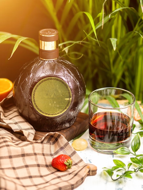 Copa de vino y botella redonda tradicional en un tablero de madera en la tabla de cocina. Con cheque mantel, frutas y hierbas alrededor.