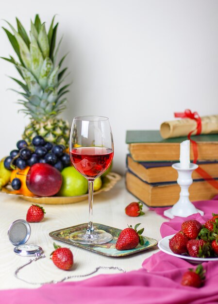 Copa de vino rosado en la mesa de madera blanca con libros antiguos y reloj, diferentes frutas tropicales y fresas