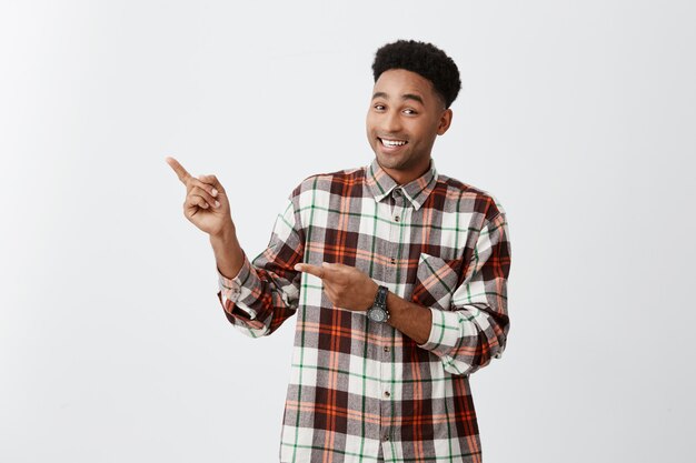 Copia espacio Chico alegre africano de piel oscura con peinado afro en camisa a cuadros apuntando a un lado con las manos en la pared blanca con expresión feliz y emocionada