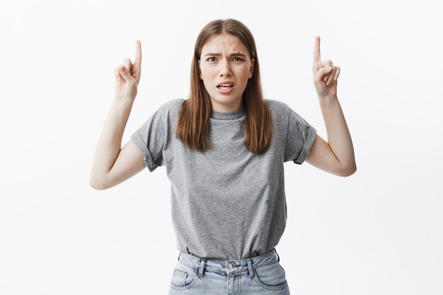 Copia espacio La emoción de las personas. Graciosa guapa morena estudiante europea en camiseta gris y jeans con expresión de disgusto