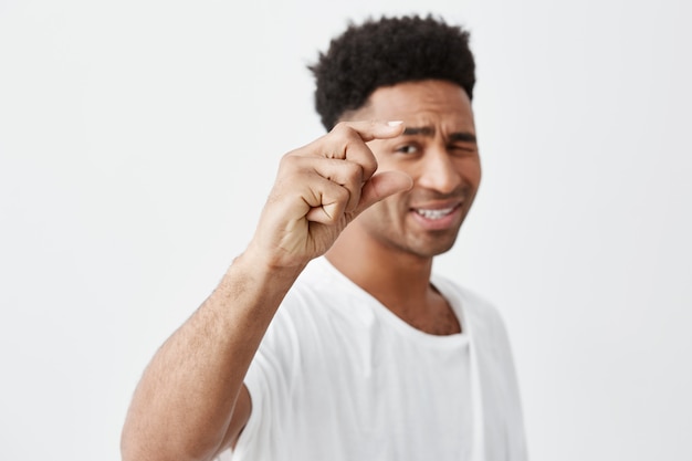 Foto gratuita copia espacio enfoque selectivo. cerca del joven y hermoso hombre de piel oscura con cabello afro en camiseta blanca gesticulando con la mano, mirando a la cámara con expresión sarcástica.