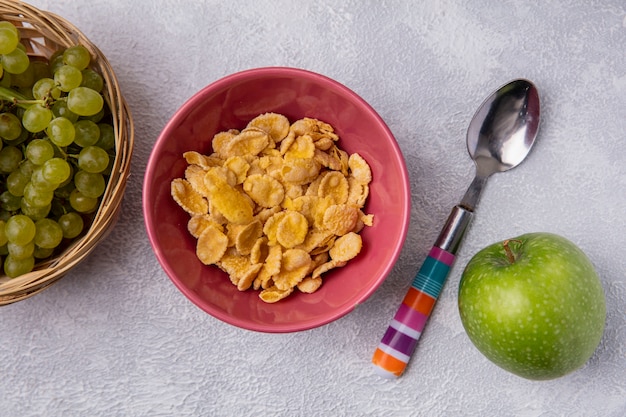 Foto gratuita copos de maíz de vista superior en un recipiente con una cucharadita con una manzana verde y uvas verdes sobre un fondo blanco.