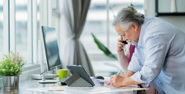 Cópula senior asiática que trabaja en casa Un anciano asiático jubilado y su esposa usan el teléfono inteligente para discutir con el cliente y verificar la información de datos con una computadora portátil en la mesa en la sala de estar en casa