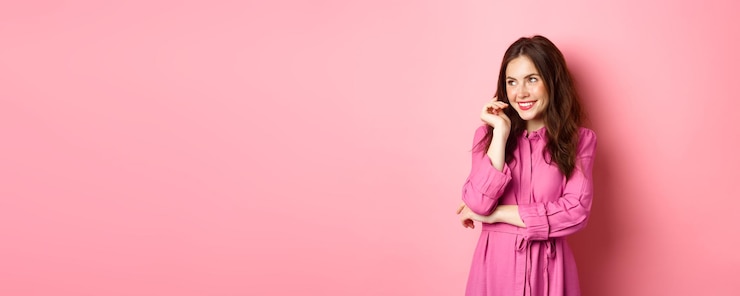 Foto gratuito coqueta mujer caucásica con bonito peinado y vestido primaveral mirando a un lado a la izquierda copyspace sonriendo pensativo de pie sobre fondo rosa