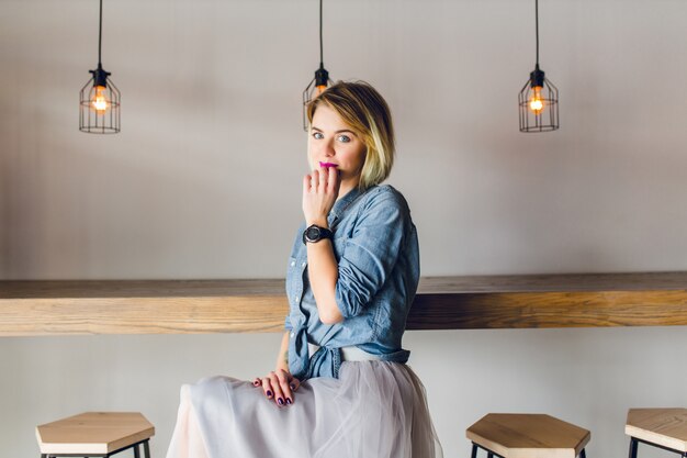 Coqueta mujer rubia con ojos azules y labios de color rosa brillante sentado en una cafetería en una silla tomando café