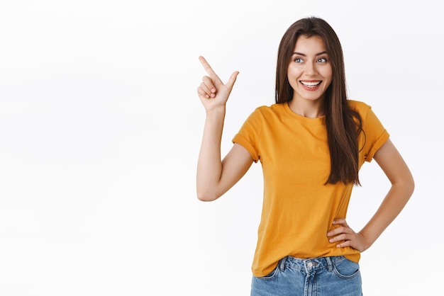 Coqueta y tonta mujer feliz con camiseta amarilla que se ríe emocionada y divertida al ver la increíble promoción del evento que apunta a la esquina superior izquierda se ve curiosa y complacida con el fondo blanco