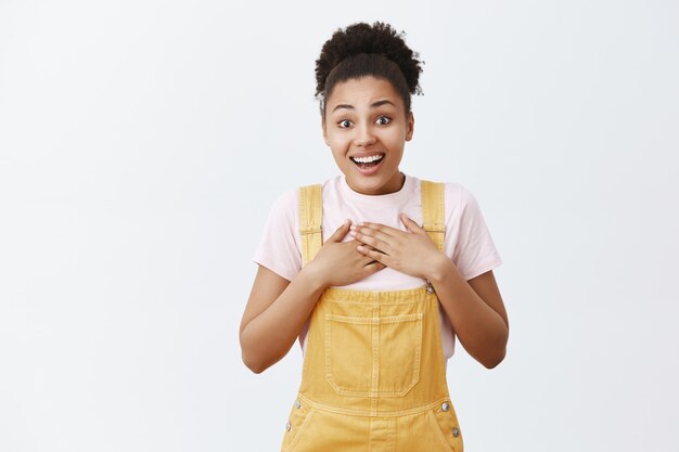 El corazón se derrite de asombrosa sorpresa. Conmovida y sorprendida mujer afroamericana de buen aspecto con un mono amarillo, tocando el pecho y sonriendo con entusiasmo, impresionada por la buena acción