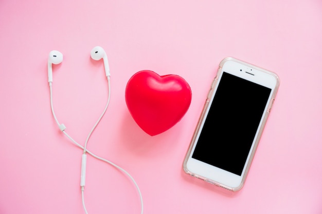 Corazón rojo entre el auricular y el teléfono inteligente en fondo rosado