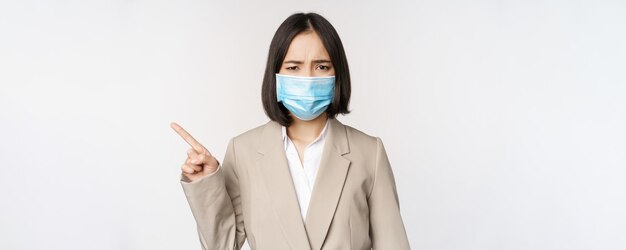 Coronavirus y concepto de trabajo Mujer de negocios asiática frustrada con mascarilla médica apuntando con el dedo a la izquierda y quejándose de pie sobre fondo blanco