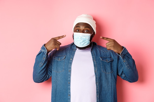 Coronavirus, estilo de vida y concepto de distanciamiento social. Feliz el hombre negro en beanie apuntando a su mascarilla, sonriendo a la cámara, de pie sobre fondo rosa