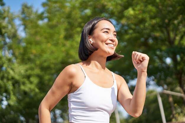 Corredor asiático sonriente de la muchacha de la aptitud en los auriculares inalámbricos que se ejecutan en el entrenamiento de jogging del parque al aire libre