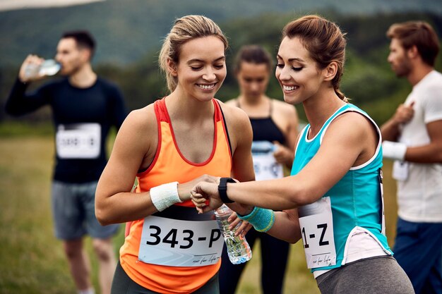 Corredor de maratón feliz mostrando reloj inteligente a su amiga antes de la carrera en la naturaleza