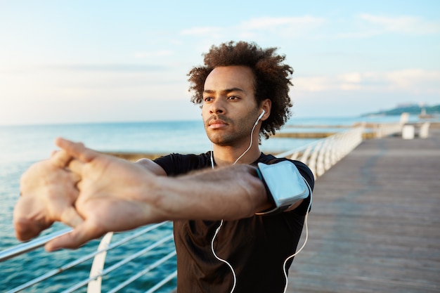 Corredor masculino afroamericano con hermoso cuerpo atlético y cabello tupido que estira los músculos, levanta los brazos mientras calienta antes de la sesión de entrenamiento de la mañana.