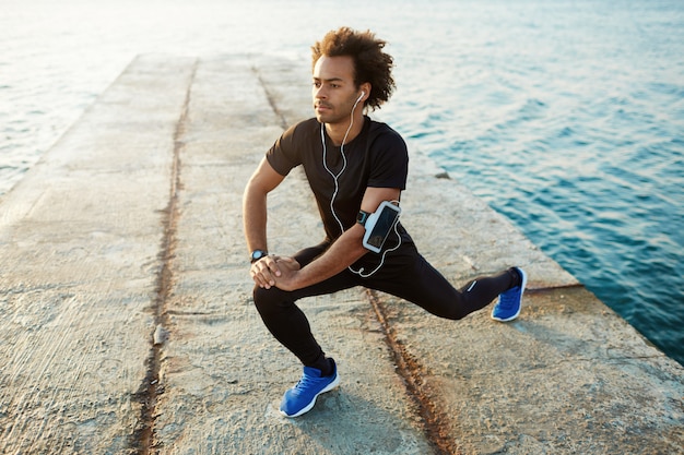 Corredor de piel oscura masculino joven con hermoso cuerpo en forma calentando sus músculos antes de entrenamiento de fuerza cardio.