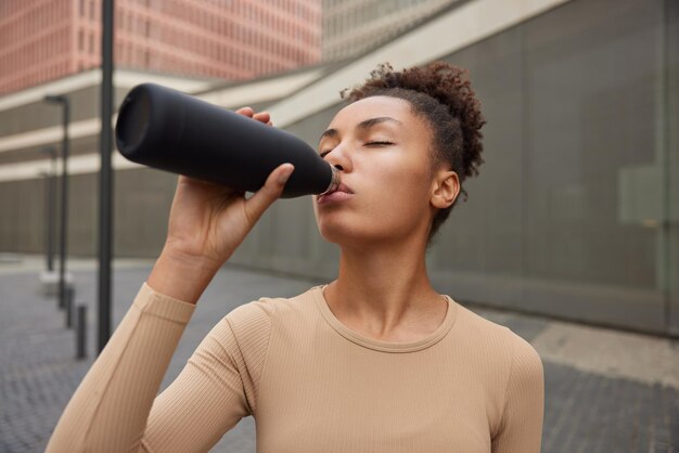 La corredora con el pelo rizado se siente sedienta después de un ejercicio intensivo bebe agua fresca de una botella vestida con ropa deportiva posa al aire libre contra un fondo borroso Atleta femenina después del entrenamiento