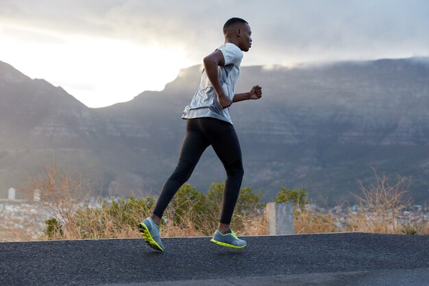 Corriendo a un joven en ropa deportiva, hace ejercicio para trotar, practica resistencia, disfruta del aire fresco cerca de las montañas. Concepto de fitness, movimiento y estilo de vida saludable. Increíble cielo azul claro durante la mañana.