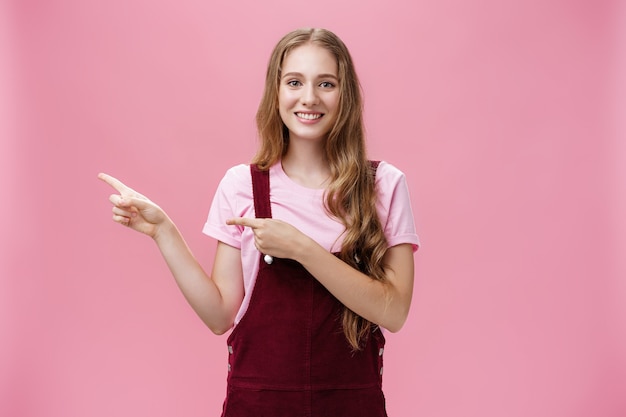 Corta y carismática jovencita con cabello largo y rubio con un mono de pana de moda apuntando a la izquierda como si estuviera invitando o mostrando algo interesante sonriendo amigablemente a la cámara sobre fondo rosa.