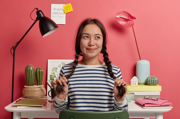 Cosas de chica hipster pensativa sobre ideas para trabajos de investigación, se sienta en el espacio de trabajo compartido contra el escritorio con cuadernos, carpetas y libros