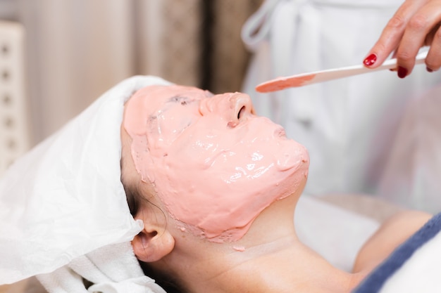 La cosmetóloga aplica la máscara de alginato con una espátula en el rostro de la mujer.