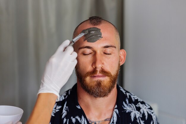 Cosmetóloga aplicar mascarilla de arcilla con pincel en la cara del joven guapo en la clínica de belleza