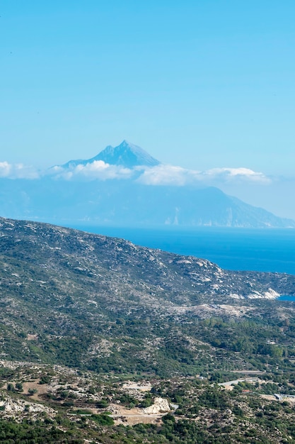 Costa del mar Egeo con colinas llenas de vegetación, edificios cerca de la costa con alta montaña llegando a las nubes Grecia