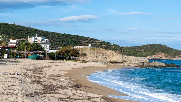 Costa del mar Egeo con edificios a la izquierda, rocas, arbustos y árboles, agua azul con olas en Sarti, Grecia