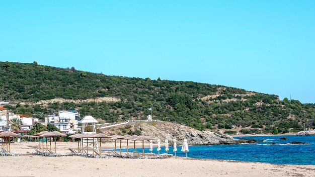 Costa del mar Egeo con edificios a la izquierda, rocas, sombrillas con hamacas, arbustos y árboles, agua azul con colina en Sarti, Grecia