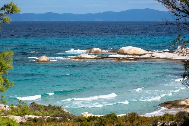Costa del mar Egeo con vegetación alrededor, rocas y árboles, agua azul con olas, Grecia