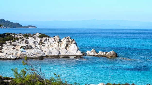 Costa del mar Egeo con vegetación alrededor, rocas y arbustos, agua azul y gente descansando, Grecia