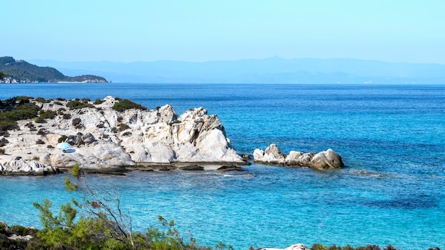 Costa del mar Egeo con vegetación alrededor, rocas y arbustos, agua azul y gente descansando, Grecia