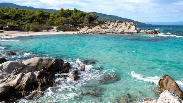 Costa del mar Egeo con vegetación alrededor, rocas, arbustos y árboles, agua azul con olas, Grecia