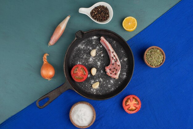 Costilla cruda en sartén negra con verduras y especias en mesa azul.