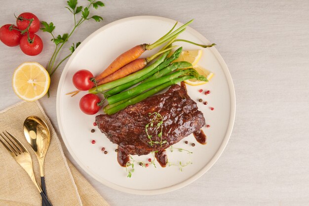 Foto gratuita costillas de cerdo asadas a la parrilla de una barbacoa de verano servidas con verduras, espárragos, zanahorias baby, tomates frescos y especias. costillas ahumadas en plato blanco sobre superficie de piedra. vista superior.