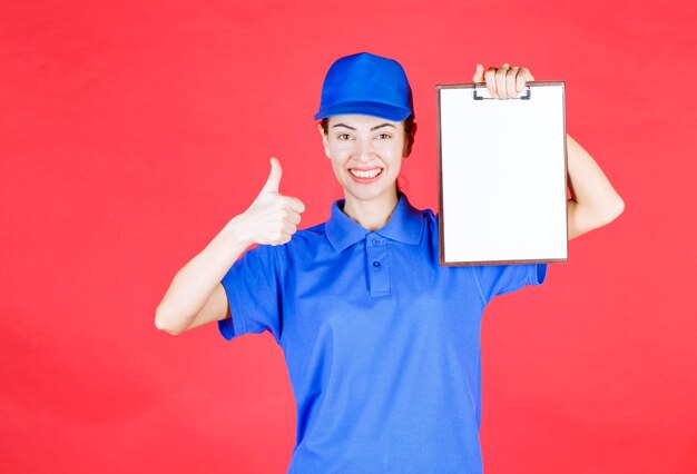 Courier girl en uniforme azul sosteniendo una lista de tareas y mostrando un signo de disfrute.